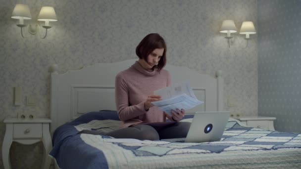 Mujer morena en vestido sosteniendo y leyendo documentos de papel sentado en la cama. Mujer estudiando las facturas y escribiendo en el portátil . — Vídeo de stock