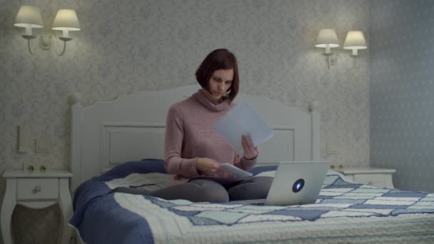 Mulher morena em vestido segurando e lendo documentos de papel sentado na cama. Feminino estudando contas e digitando no laptop . — Vídeo de Stock