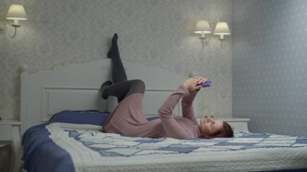 穿着网上冲浪衣服的布鲁内特女人躺在床上用智能手机。 女尸躺在后座上，腿缓缓地靠在床头板上 — 图库视频影像