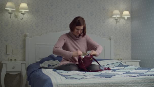 Молодая брюнетка ищет телефон в маленькой сумке на кровати. Женщина трясет сумку и находит гаджет в беспорядке и падает в постель — стоковое видео