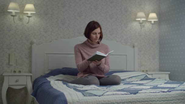 Брюнетка в платье читает бумажную книгу, сидя на кровати. Женщина переворачивает страницы в бумажной книге в замедленной съемке . — стоковое видео