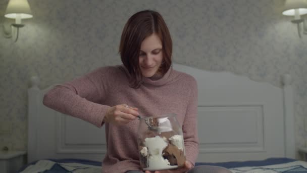 Mladá brunetka si užívá jíst vanilku a čokoládový kbelík se zmrzlinou v rukou. Žena ráda poruší dietu sladkostmi na posteli. — Stock video