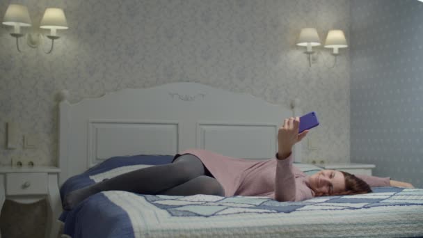 Femme brune endormie en robe couchée sur le lit et mettant le smartphone de côté. Femme se réveillant, regardant l'heure au téléphone et tombant pour se détendre . — Video