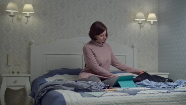 Mujer joven mirando su ropa sentada en la cama para venderla en línea. Vestido de mujer sosteniendo en percha y tomando fotos para su aplicación . — Vídeo de stock