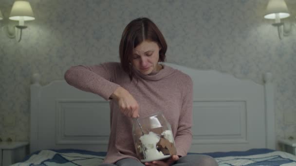 Jeune femme brune en pleurs mangeant un seau de vanille et de crème glacée au chocolat tenant dans les mains assises sur le lit. Femmes énervées mangeant des bonbons . — Video