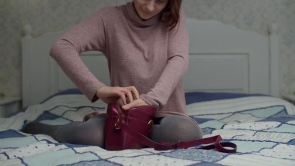 Młoda brunetka szukająca telefonu w małej torbie na łóżku. Kobieta potrząsa torbą i znajduje gadżet w bałaganie. — Wideo stockowe