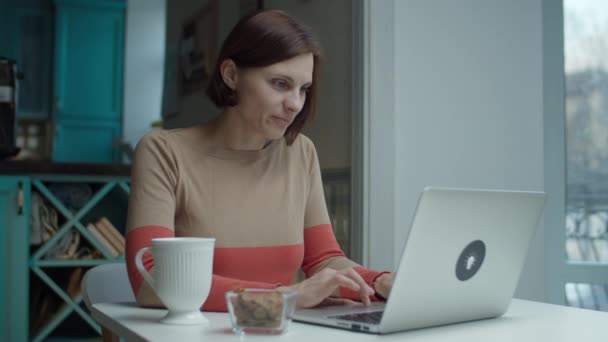 Mujer joven que trabaja en el ordenador portátil sentado en la mesa con aperitivo y una taza de bebida caliente. Mujer bebiendo café y sonriendo a gadget . — Vídeo de stock