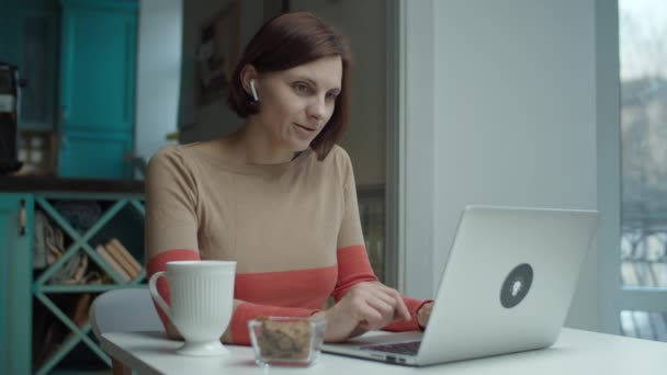 젊은 여성 이 책상에 앉아 무선 이어폰을 끼고 노트북을 사용하여 온라인으로 이야기하고 있습니다. 컴퓨터 일을 하는 여성의 이야기. — 비디오