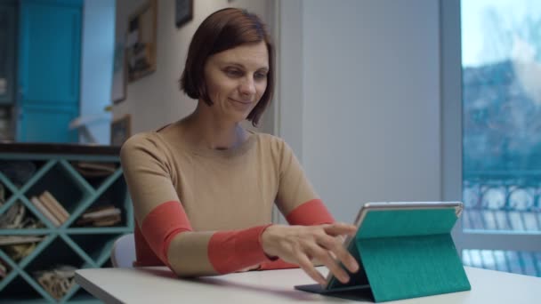 Brünette Frau surft online auf Tablet-Computer sitzt am Tisch. Frauen berühren Gadget-Bildschirm und lächeln in Zeitlupe — Stockvideo