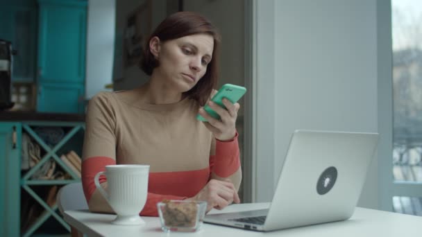 Young woman sitting at desk, distracting with phone while working with laptop. Female using cell instead of working on computer. — ストック動画