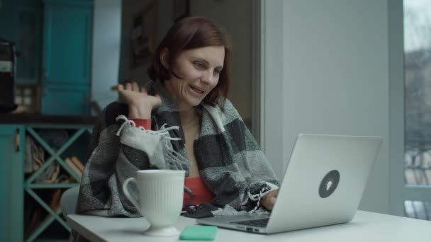 Mujer joven en manta sentada en el escritorio y haciendo videollamadas con computadora portátil. Mujer bebiendo café o té y hablando en línea en la computadora — Vídeos de Stock