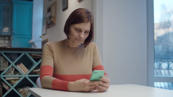 Mujer morena surfeando en línea en el teléfono inteligente sentado a la mesa. Pantalla de gadget táctil femenina y sonriendo en cámara lenta — Vídeo de stock