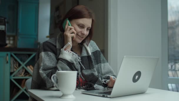Ung kvinna i filt sitter vid skrivbordet distrahera med mobilsamtal medan du använder laptop. Kvinna pratar per telefon och arbetar på dator — Stockvideo