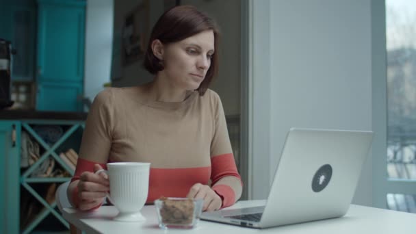 Ung kvinna som arbetar på laptop sitter vid bordet med mellanmål och kopp varm dryck. Kvinnligt drickande kaffe och leende att gadget. — Stockvideo