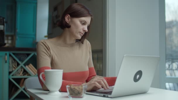 Jonge vrouw zit achter de balie, neemt draadloze oortjes aan en praat online met een laptop. Vrouw praten tijdens het werken op de computer. — Stockvideo