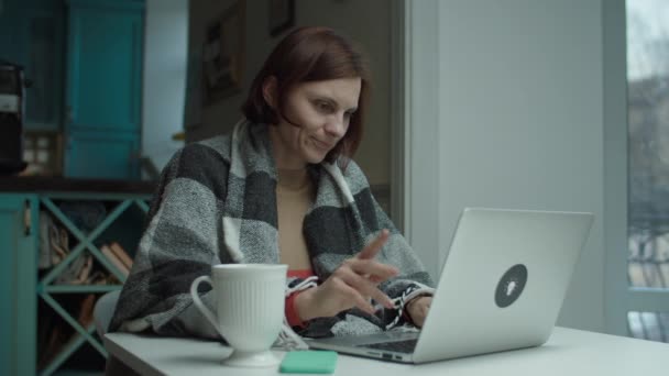 Young woman in blanket sitting at desk distracting with cell call while using laptop. Female talking by phone and working on computer — Stock Video