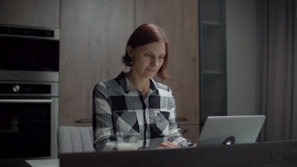 Donna freelance con computer portatile seduto al tavolo della cucina. Donna che beve latte con snack. Donna che lavora con gadget a casa . — Video Stock