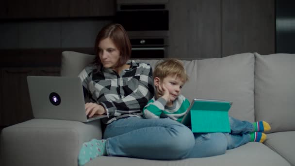 Madre trabajadora con portátil e hijo con tableta sentada en un acogedor sofá. Familia usando gadgets phubbing entre sí . — Vídeo de stock