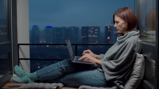 Die freiberufliche Mitarbeiterin sitzt am Laptop auf der gemütlichen Fensterbank mit Blick auf die Stadt am Abend. hohe Gebäude mit Lichtern in grauer Stadtkulisse. — Stockvideo