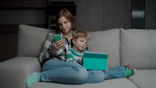 Мать со смартфоном обнимает сына с планшетным компьютером, сидящим на уютном диване. Семья, использующая гаджеты, дублирующие друг друга . — стоковое видео