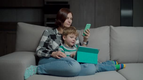 Madre con teléfono inteligente abrazando hijo con la tableta de ordenador sentado en el sofá acogedor. Familia usando gadgets phubbing entre sí . — Vídeo de stock