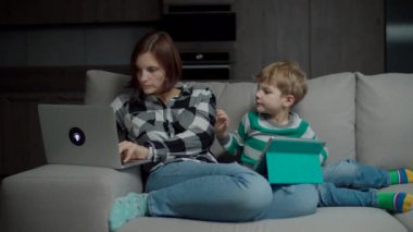 Bilgisayarlı çalışan bir anne ve rahat koltukta oturan tabletli bir oğlu var. Aile, birbirlerini kandıran aletler kullanıyor.. 