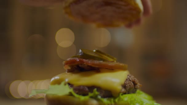 Man hand afwerking koken zelfgemaakte hamburger door te drukken op top broodje. Roterende hamburger met vleessnijder, gesmolten kaas, tomaten, komkommers en saladebladeren. — Stockvideo