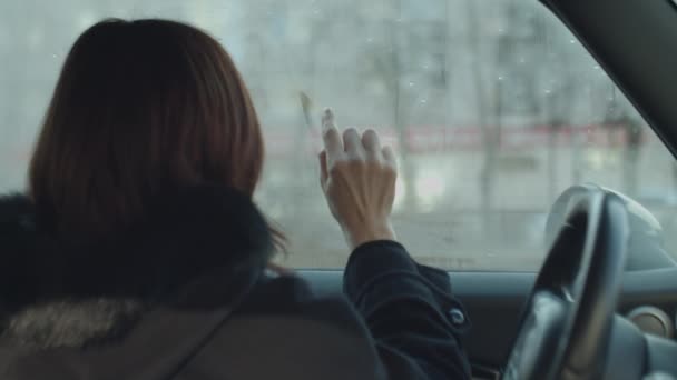 Back of brunette woman writing sign MINI by finger on misted car glass. Finger painting on misted window in car. — Stock Video