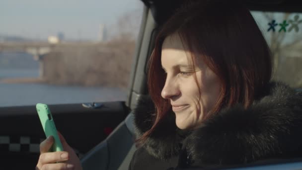 Mujer adulta de 30 años sentada en coche y viendo contenido divertido en línea en el teléfono inteligente en las manos con el paisaje soleado del río de la ciudad . — Vídeo de stock