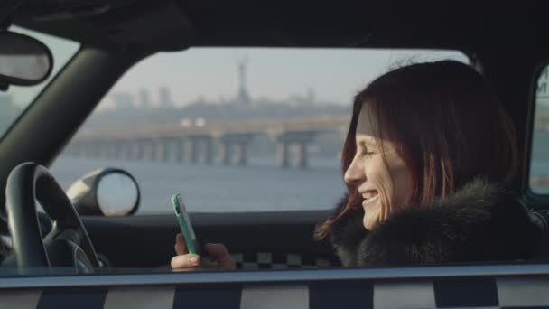 Mujer adulta de 30 años sentada en coche y viendo contenido divertido en línea en el teléfono inteligente en las manos con el paisaje soleado del río de la ciudad . — Vídeos de Stock