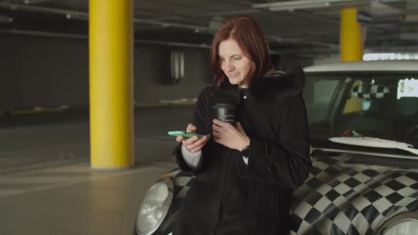 Mujer de 30 años adulta en la capucha del coche surfeando en línea a través de smartphone con taza de café reutilizable en la mano en la zona de aparcamiento. Mujer revisando línea de tiempo de redes sociales . — Vídeo de stock