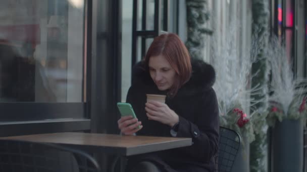 都会のカフェに座って熱いコーヒーを楽しむセルを持つ30代の大人の女性。女性ともにお茶を飲みますガジェット画面に座ってカフェ. — ストック動画