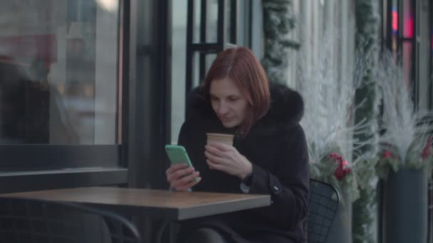 Mujer adulta de 30 años con celda disfrutando de café caliente sentada en el café urbano. Mujer con taza de té mirando a pantalla gadget sentado en la cafetería . — Vídeo de stock