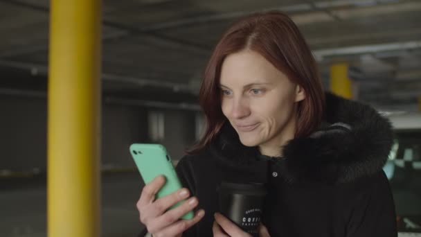 Mujer de 30 años adulta en la capucha del coche surfeando en línea a través de smartphone con taza de café reutilizable en la mano en la zona de aparcamiento. Mujer revisando línea de tiempo de redes sociales . — Vídeos de Stock