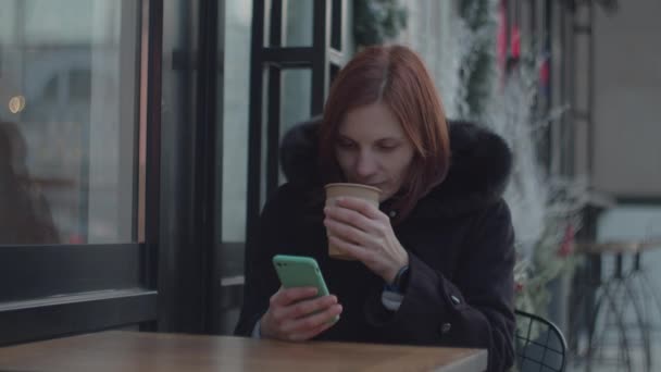 30 років доросла жінка з клітиною насолоджується гарячою кавою, сидячи в міському кафе. Жінка з чашкою чаю дивиться на екран гаджета, сидячи в кафе . — стокове відео