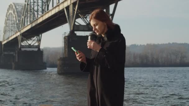 30s femme adulte profiter du café chaud du matin avec smartphone dans les mains. Température hivernale froide et venteuse sous le pont fluvial . — Video