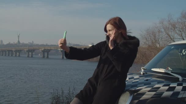 Female 30s adult in black coat on car hood making online selfie video to social media with fall city river landscape. — 비디오