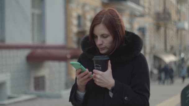 Frozen 30s adult woman enjoying hot coffee cup with smartphone in cold hands. Cold windy winter weather on city streets. — Stock Video