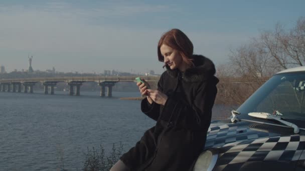 Laughing female 30s adult in black coat on car hood surfing online via smartphone in hands with fall city river landscape. — 비디오
