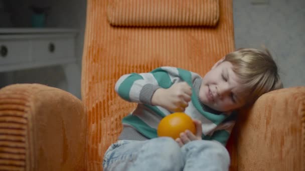 Blonde preschool boy trying to peel an orange sitting in cozy orange armchair. Kid kicking orange because cant cope with peeling fruit. — 비디오