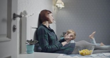 Anne ve oğlu evdeki yatakta televizyon izlerken eğleniyorlar. Anne cips yiyor, oğul uzaktan kumandayı çekiyor.. 