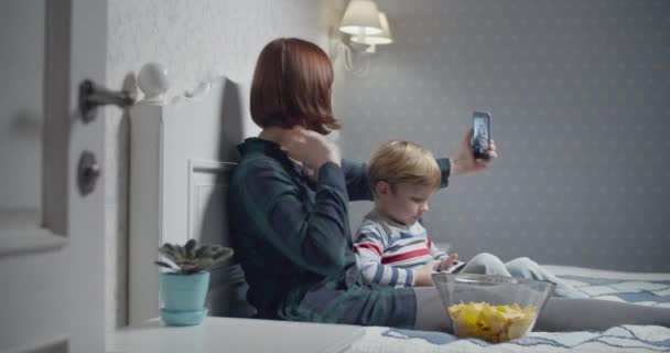 Young mother recording selfie video by smartphone and son playing video games sitting on bed at home. Family evening pastime. Boy winning in video game and rising up hands — Stock Video