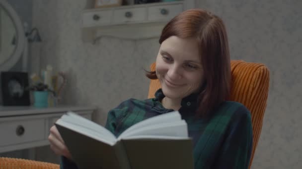 Jonge vrouw die papieren boek leest zittend in een gezellige oranje fauteuil in de slaapkamer. Vrouwelijke ruiken en draaien pagina 's in boek in slow motion. — Stockvideo