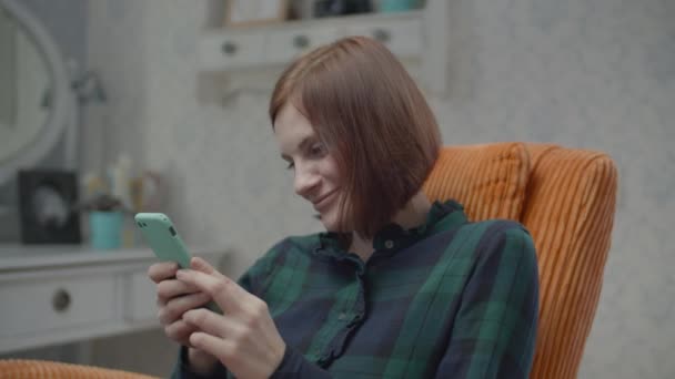 Junge weibliche brünette Erwachsene surfen online im Smartphone sitzt in gemütlichen orangefarbenen Sessel im Schlafzimmer in Zeitlupe. — Stockvideo