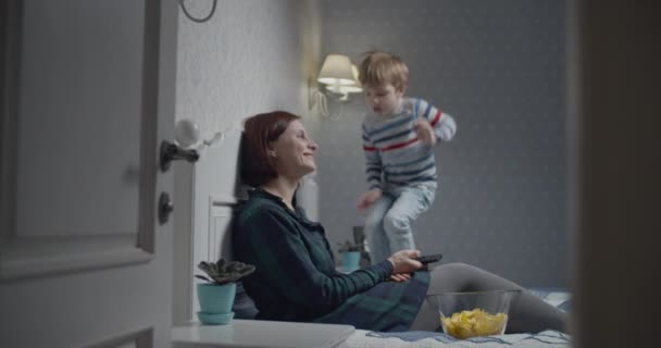Mor och son har kul medan de tittar på TV på sängen hemma. Ungen hoppar i säng medan mamma tittar på tv. — Stockvideo