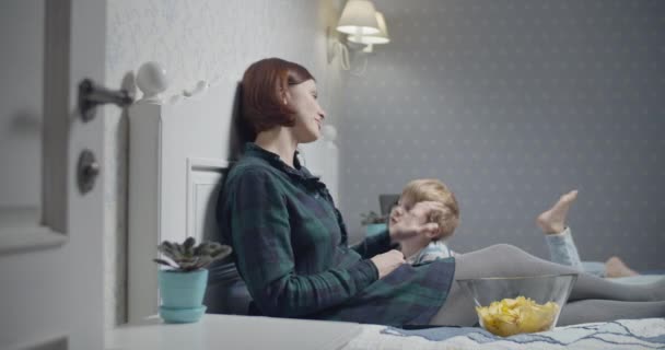 Mor och son har kul medan de tittar på TV på sängen hemma. Mamma äter chips, son drar tillbaka fjärrkontrollen. — Stockvideo