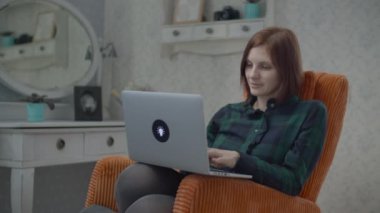 Yatak odasındaki rahat turuncu koltukta dizüstü bilgisayarda çalışan serbest bir kadın. Evde aletlerle çalışan bir kadın..