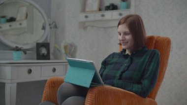 Genç kadın esmer yetişkin yatak odasındaki rahat turuncu koltukta oturup tablet bilgisayardan içeriği izliyor. Kadın yüzüne yansıyan Gadget ekranı. 