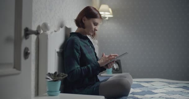Молодая брюнетка взрослая работает за планшетным компьютером, сидя на кровати. Женщина трогает экран гаджета пальцами . — стоковое видео