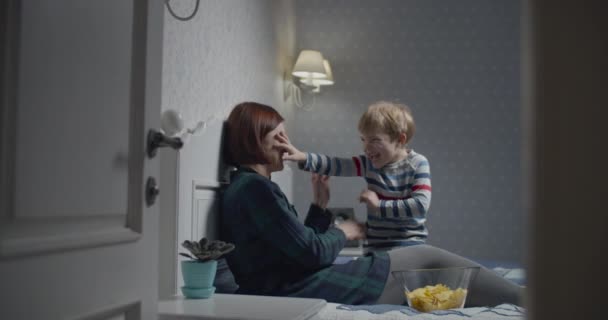 Matka a syn se baví při sledování televize doma na posteli. Dítě zavírá máminy oči a směje se. Šťastný rodinný čas. — Stock video